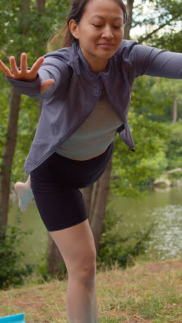 Video-Vertical-De-Una-Mujer-Vestida-Con-Ropa-Deportiva-Haciendo-Yoga,-Estirándose-Y-Equilibrándose-En-Un-Bosque-Junto-A-Un-Lago-O-Río,-Disfrutando-De-La-Paz-Y-La-Belleza-De-La-Naturaleza,-Filmado-En-Tiempo-Real